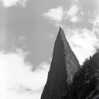 Vygenerováno pomocí umělé inteligence DALLE-2 dotazem: Old black and white photo of a very thin and high mountain which looks like a needle reaching skies. Tmavý obrys úzké hory, trojúhelníkového charakteru, sahající až do oblak
